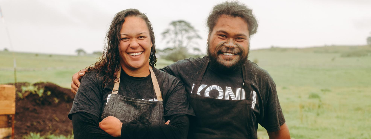 Two beautiul Māori people with arms around shoulders wearing black Konei branded shirts, background is a rural setting