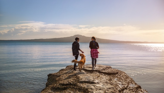 Two women and a dog standing on a rock at Kohimaramara beach Auckland looking at Rangitoto Island